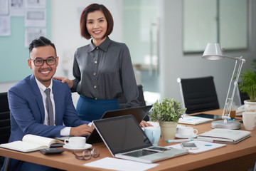 Portrait of Asian young businessman sitting at office desk typing on laptop with beautiful businesswoman standing near by him, they smiling at camera at office