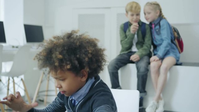 Two Caucasian kids bullying mixed race schoolboy typing on laptop computer at desk in elementary school classroom. Students pointing at classmate and discussing in intolerant way behind his back,