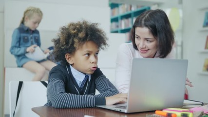 Rack focus of blonde schoolgirl reading book while teacher explaining task on laptop computer to his male classmate sitting at desk in elementary school classroom