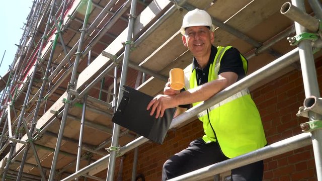 Male builder foreman, surveyor, worker or architect working on construction building site standing on scaffolding with a clipboard and drinking mug of tea or coffee
