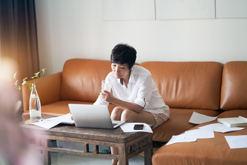 Fototapeta na wymiar Asian self employed woman sitting on couch & working on laptop at home