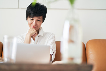 confident Asian self employed woman sitting on couch smiling & working on laptop at home