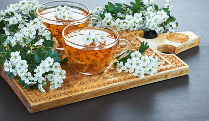 Haw thorn flower tea, two cups of healthy homemade infusion drink with blossom and branches of a tree on cutting board on dark background, close-up, copy space for your design, overhead view