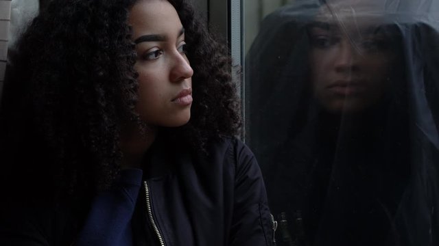 Sad depressed beautiful female mixed race African American girl teenager young woman teen wearing a black jacket sitting looking out of a window 