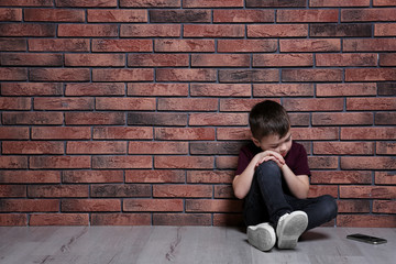 Sad little boy with mobile phone sitting on floor near brick wall. Space for text