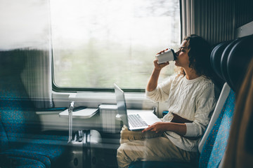 Freelancer girl in white sweater working with laptop in the train and drinking coffee , business...