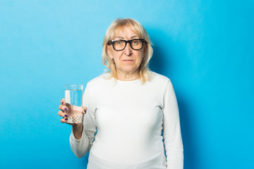 Old lady with glasses holding a glass of water against a blue background. Concept of thirst, dehydration