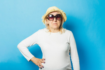 Old woman in a straw hat and sunglasses on a blue background. The concept of the onset of summer,...