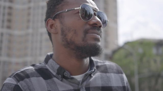 Close up face of confidient African American man standing against the background of urban building squinting in the sun, then putting on sunglasses close-up. The guy resting outdoors alone, skyscraper
