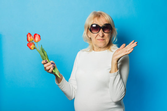 Old woman in sunglasses is holding a bouquet of tulips on a blue background. Concept of the onset of spring, summer time, vacation, camping