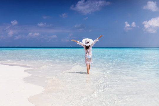 A woman in white summer dress an sunhat stands in endless turquoise sea on a sunny day and enjoys her tropical vacation