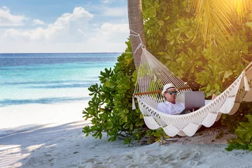  Arbeiten im Urlaub: Geschäftsmann liegt mit Laptop auf einer Hängematte unter Palmen und arbeitet © moofushi