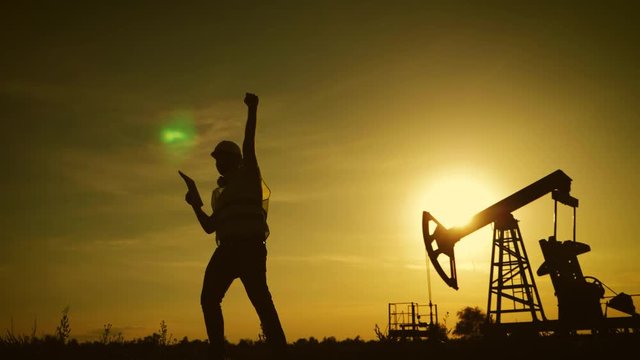 Silhouette of happy oilfield worker at crude oil pump in the oilfield at golden sunset. Industry, oilfield, people and development concept.