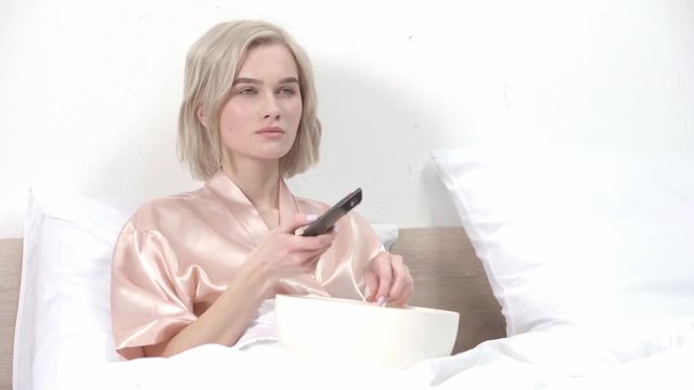 attractive blonde girl eating popcorn while holding remote controller and watching movie in bed 