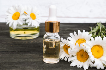 Chamomile essential oil in a glass bottle with a pipette is on the table near the chamomile flowers.