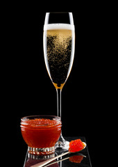 Elegant glass of yellow champagne with red caviar on golden spoon and glass container of caviar on marble board on black background.