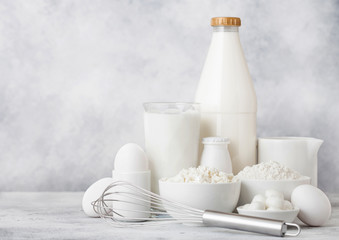 Fresh dairy products on white table background. Plastic bottle and glass of milk, bowl of cottage cheese and baking flour and mozzarella. Eggs and cheese. Steel whisk.