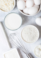 Fresh dairy products on white table background. Glass of milk, bowl of flour, sour cream and cottage cheese and eggs. Steel whisk. Top view.