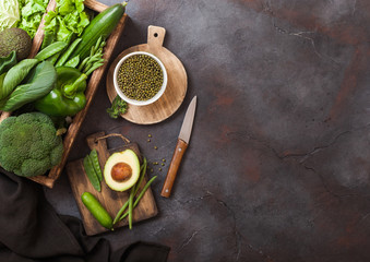 Assorted green toned raw organic vegetables in wooden box on dark background. Avocado, cabbage, cauliflower and cucumber with trimmed and mung beans and chopping board with knife. Space for text