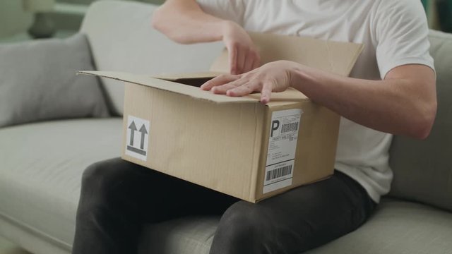 A young man opens a cardboard parcel on the couch in his living room