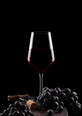Glass of red wine with dark grapes and vintage corkscrew opener and cork on wooden board on black background.