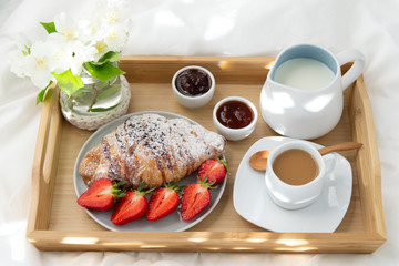 Breakfast in bed. Wooden tray with coffee , jam, strawberries and croissants. Decoration of delicate white flowers. Beautiful sunlight.