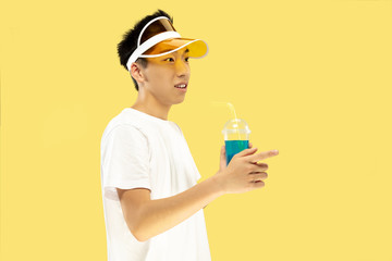 Korean young man's half-length portrait on yellow studio background. Male model in white shirt and yellow cap. Drinking cocktail. Concept of human emotions, expression, summertime, vacation, weekend.