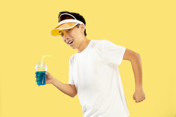 Korean young man's half-length portrait on yellow studio background. Male model in white shirt and yellow cap. Drinking cocktail. Concept of human emotions, expression, summertime, vacation, weekend.
