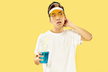 Korean young man's half-length portrait on yellow studio background. Male model in white shirt and yellow cap. Drinking cocktail. Concept of human emotions, expression, summertime, vacation, weekend.