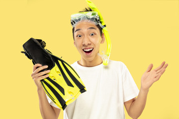 Korean young man's half-length portrait on yellow studio background. Male model in white shirt and swimglasses. Holding flippers. Concept of human emotions, expression, summertime, vacation, weekend.