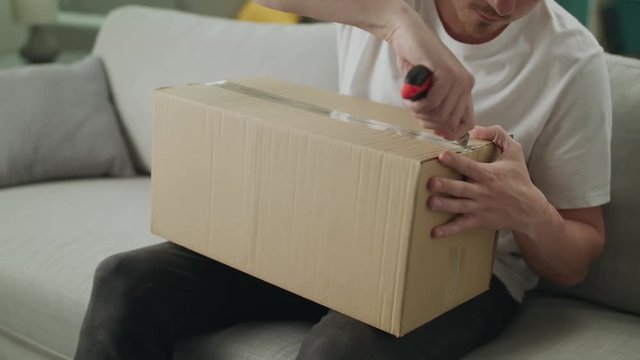 A young man opens a cardboard parcel on the couch in his living room