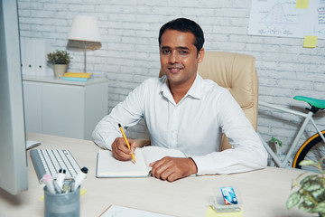Portrait of professional sitting at the table and writing his plans and ideas in notepad at modern office