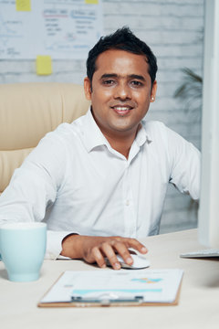 Portrait of Indian confident businessman sitting at the table using computer in his work and smiling at camera at office