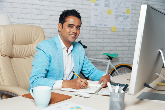 Portrait of Indian businessman in stylish suit smiling at camera while making notes in notepad at the table at office