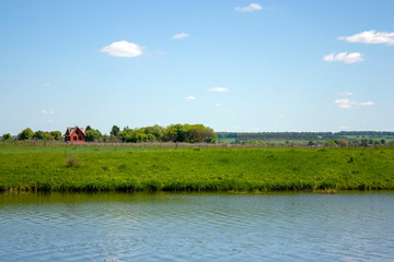 Lone red brick house among green trees. Unfinished house stands near the river. Green foliage of trees and grass by the river. Blue sky and clouds over the horizon on a sunny summer day.