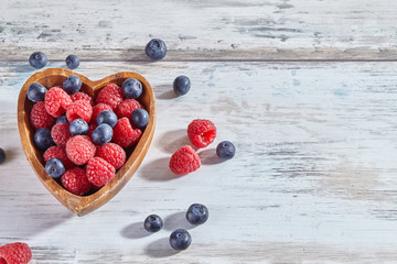 Ripe sweet raspberries and blueberries in a wooden heart-shaped bowl on white rustic wooden...
