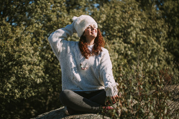 A beautiful woman is sat on a rock in the nature. She is smiling, with the sun shining her face. Her right hand is behind her head. She has long brown curly hair and is wearing a white hat.