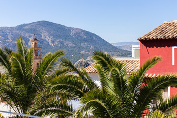 Fototapeta na wymiar Tiled roofs and dome with ceramic tiles of the church and bell tower and palm trees in the foreground against the backdrop of the mountains, Spain