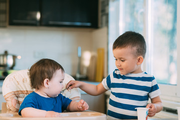 Kids in the kitchen, brother feeds baby sister yogurt with a spoon, she helps parents
