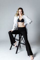 Fashion model sitting on a chair in a white blouse and flared pants barefoot on a white background