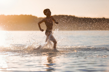 On the nice sunny summer day the small boy is playing, running, jumping at the lake. Happy moments of childhood. Reflection of the silhouette on the water. Beautiful place Svityaz lake, Ukraine.