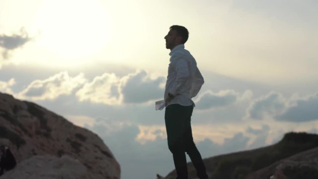 Bearded man in a white shirt standing on top of a mountain peers at the clouds. Young stylish guy stand on the high hill of mountain look far away. Cyprus. Paphos