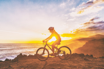 Mountain biking cyclist girl riding MTB bike on coast trail against sunset. Silhouetter of woman...
