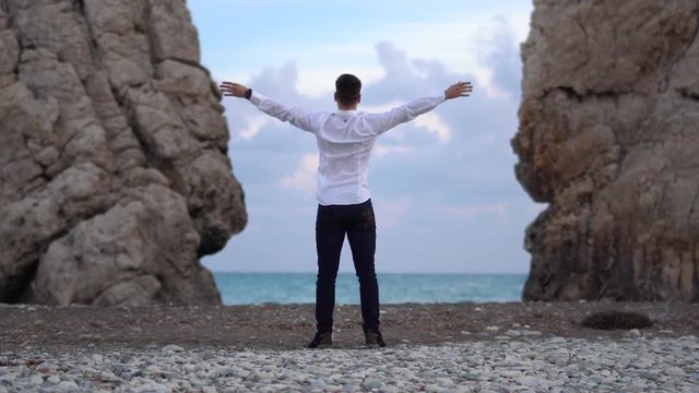 Man in a white shirt and dark jeans throwing his hands byside, enjoying freedom, believe future success. Happy tourist on rock top, victory and purpose. Cyprus. Paphos