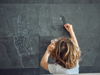 Back view of long hair boy writing by chalk multiplication table on grey stone wall
