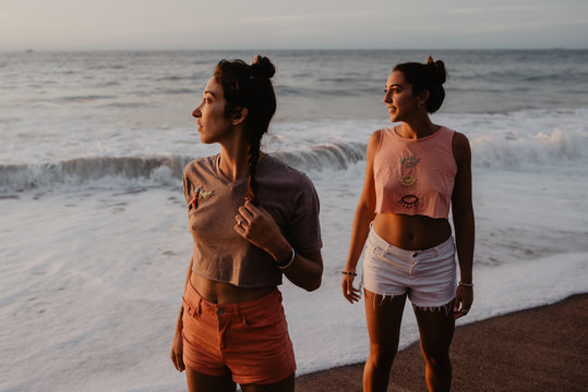 Two slim young females in shorts and bras looking away while standing on sandy shore against cloudy gray sky at sunset