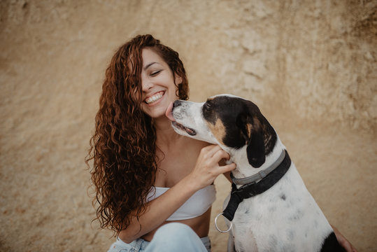 Funny dog licking cheek of excited young lady against weathered building wall on street