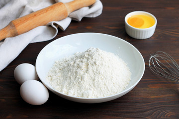 A plate with flour, eggs, a rolling pin and whisk on a white wooden background. Pancakes ingredients. Egg in the bowl.