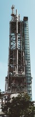 A narrow vertical view from the ground of a contemporary oil refinery building or a fuel factory facility in an industrial zone, with plenty of pipes, metal beams, and stairs; elevator on the right