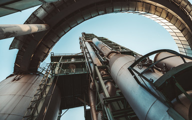 Wide-angle bottom view of a contemporary construction of an oil refinery or a modern fuel factory facility in an industrial zone, with a round bridge, plenty of pipes, iron beams, tanks, and stairs